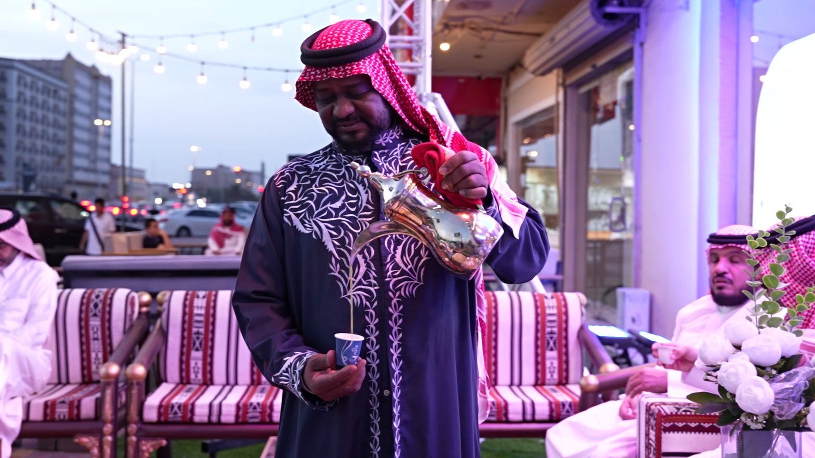 Host pouring Arabic coffee for guests at an outdoor majlis at dusk
